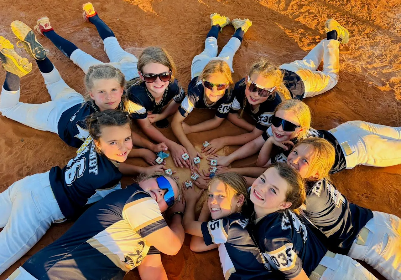 A softball team lying in a circle on the infield dirt, heads together, showing off their championship rings