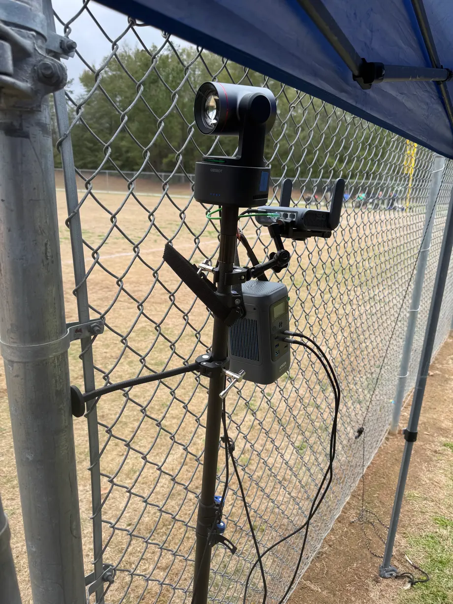 A pan-tilt-zoom camera clamped to the backstop fence, wired to a battery pack, looking out over the infield